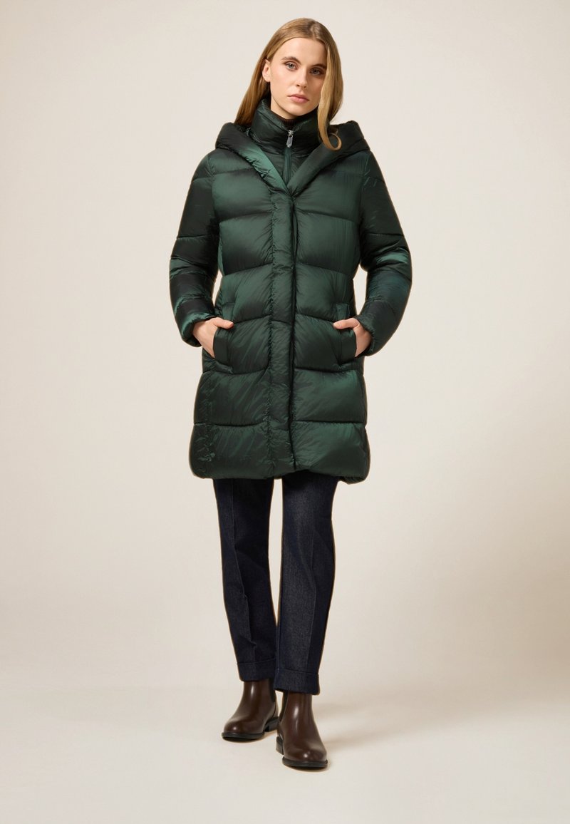 Mørkegrøn dunjakke med hætte, quiltet design, forlommer og lynlåslukning. Kombineret med mørke jeans og brune ankelstøvler.