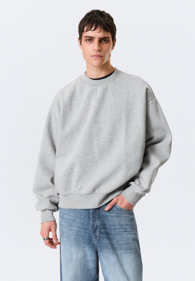 Grey sweatshirt with a round neckline, dropped shoulders, and ribbed cuffs. Worn with blue slightly distressed jeans.