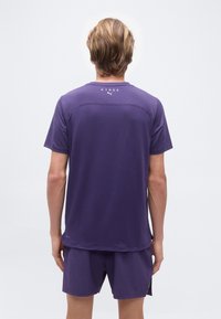 Άτομο με κοντά ξανθά μαλλιά που φοράει μωβ μπλουζάκι HYROX Puma και αντίστοιχα σορτς, στέκεται γυρισμένο πλάτη σε λευκό φόντο.