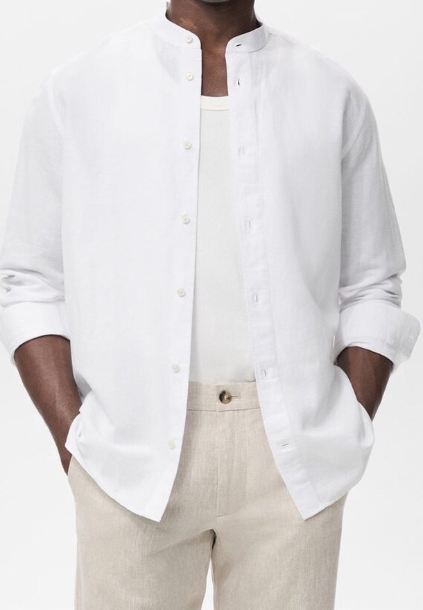 Homme portant une chemise blanche à col mandarin avec les manches retroussées, un maillot blanc en dessous et un pantalon beige, les mains dans les poches.