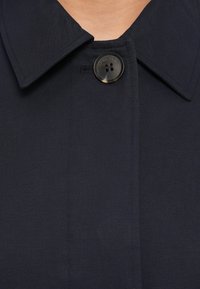 Μαύρο παλτό με κουμπιά, με φαρδύ γιακά, λεία υφή υφάσματος, εμφανή ραφή και ένα μόνο σκοτεινό κουμπί στον λαιμό.