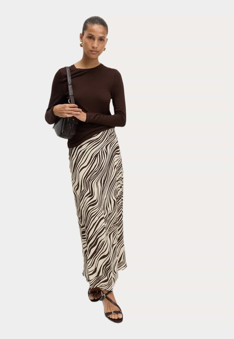 Hnědý top s dlouhým rukávem zkombinovaný s krémovo-hnědou vzorovanou maxi sukní. Doplněno černou kabelkou přes rameno a černými sandály na šněrování.