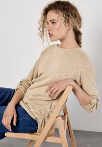 Beige långärmad topp med strukturerad finish, rundad halsringning, knappdetaljer på ärmarna, kombinerad med blå jeans, sittande på en trästol.