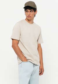 Beige cotton t-shirt with short sleeves and crew neckline, featuring a subtle logo. Paired with light blue denim jeans and a checkered cap.