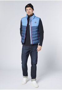 Blue puffer vest with horizontal stitching, red zipper accents, and black logo. Worn over a black long-sleeve shirt and paired with dark jeans and white sneakers.