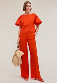 Top in cotone rosso con dettagli a cravatta sulle maniche, abbinato a pantaloni rossi a gamba larga. La modella tiene una borsa beige e indossa sandali rossi coordinati.