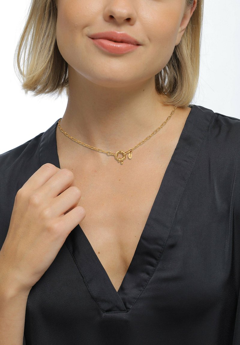 Guldkæde choker halskæde med cirkulære og ovale led, der har en lille charm. Båret over en sort satinbluse med v-hals.