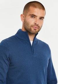 Blauer Reißverschluss-Pullover aus Strickstoff. Verfügt über einen hohen Kragen und eine gerade Passform. Glatte Textur mit gerippten Akzenten am Kragen.