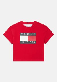 Punainen puuvillainen lyhyt t-paita, jossa on suorakulmainen grafiikka, jossa lukee "TOMMY HILFIGER" mustalla tekstillä ja kontrastiväreillä valkoisina ja punaisina lohkoina.