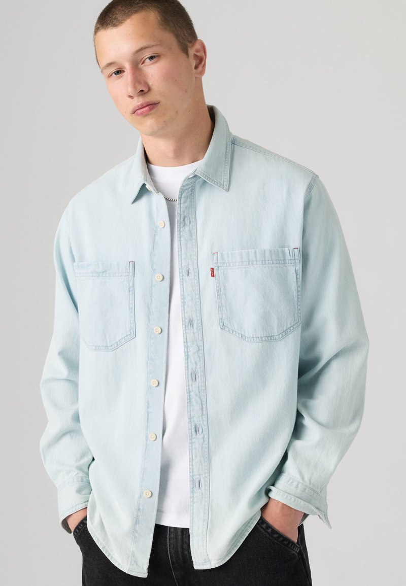 Jeune homme portant une chemise en jean bleu clair sur un t-shirt blanc, les mains dans les poches, sur un fond gris uni.