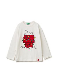 Biała koszula z długim rękawem z grafiką Snoopiego na czerwonym domku dla psa ozdobionym kolorowymi światełkami bożonarodzeniowymi. Czerwona metka przy dolnym brzegu.