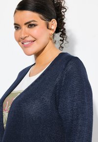Marineblauer Strickcardigan mit strukturiertem Muster, offener Vorderseite und weiten Ärmeln, getragen über einem weißen Hemd mit grünem Grafikdruck.