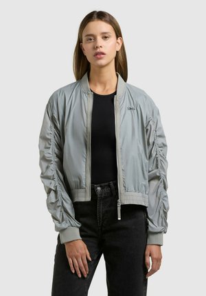 Jeune femme portant une veste bomber grise claire avec des manches froncées, un t-shirt noir et un jean noir taille haute, debout devant un fond uni.