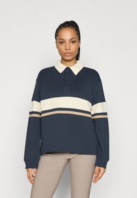 Granatowa bluza polo z długimi rękawami, kremowym kołnierzem oraz poziomymi paskami w kolorze kremowym i beżowym na wysokości klatki piersiowej.