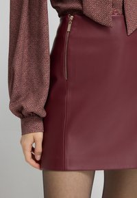 Krótka burgundowa spódnica z ekoskóry z bocznym zamkiem błyskawicznym. Tkanina ma gładką teksturę. Połączona z wzorzystą bluzką z długim rękawem.