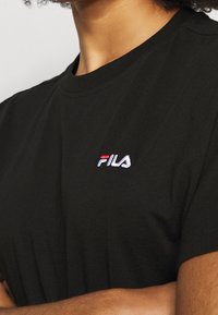 Svart bomullströja med rund hals och korta ärmar, pryds av en liten broderad vit och röd FILA-logotyp på vänster bröst.