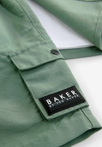 Zelená texturovaná bunda; detailní záběr na kapsu s černou etiketou "BAKER BY TED BAKER." Obsahuje detail tmavého knoflíku.