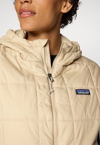 Šviesiai rusvas pūkinis švarkas su kapišonu, turintis priekinį užtrauktuką, siuvinėtą tekstūrą ir mėlynai juodą "Patagonia" logotipą kairiajame krūtinės šone.
