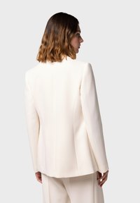 Blazer su misura color crema, design strutturato, tessuto liscio, colletto affilato, spacco singolo sul retro e dettagli delle cuciture sottili.