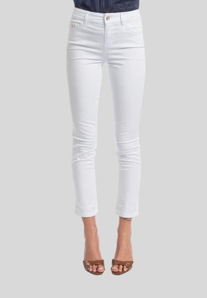 Pantaloni bianchi aderenti alla caviglia indossati con tacchi marroni a punta aperta su uno sfondo semplice.