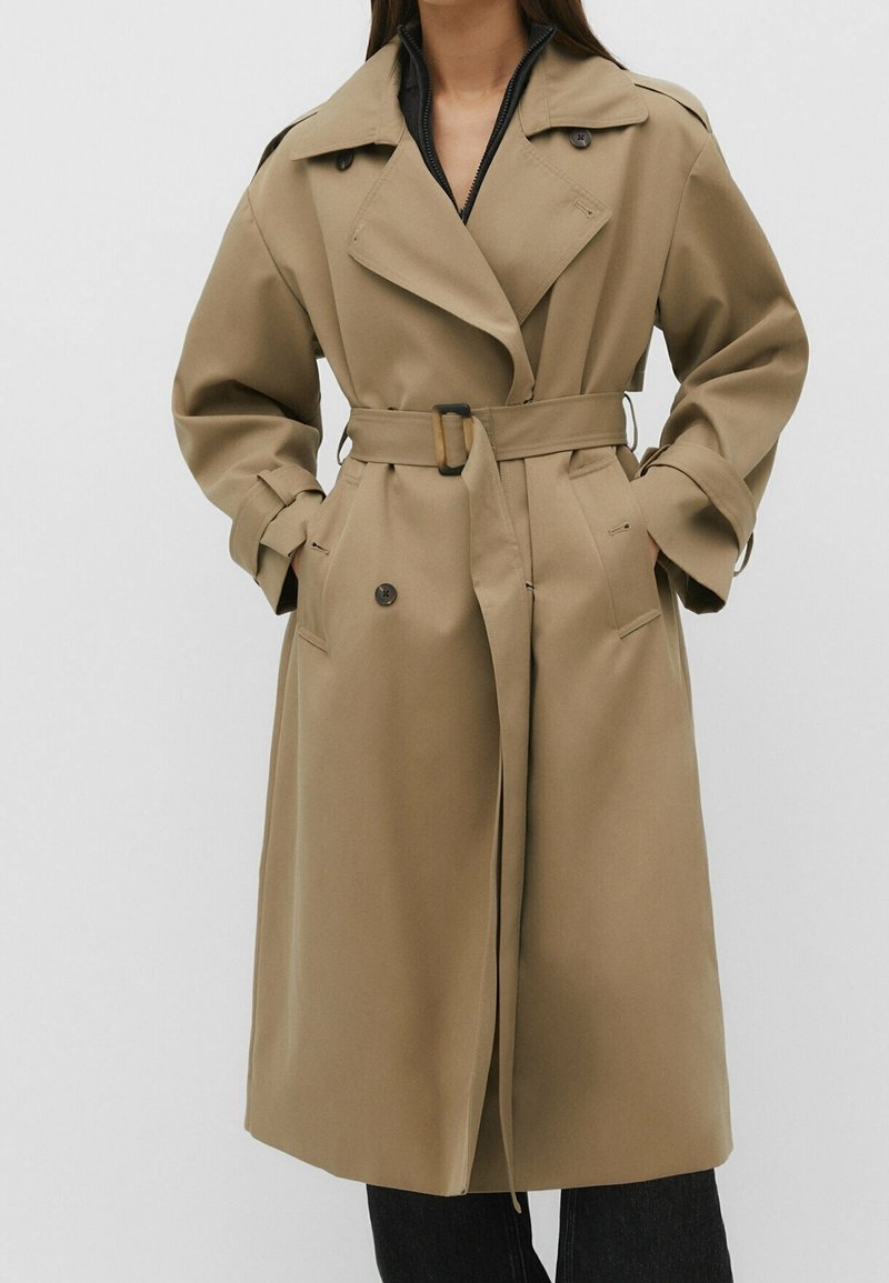 Femme portant un trench-coat beige ceinturé avec de larges revers et des détails de boutons, debout les mains dans les poches devant un fond uni.
