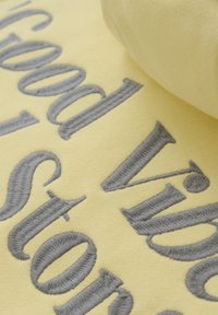 Tissu jaune avec un texte brodé gris, présentant une texture douce et un bord roulé visible dans le coin supérieur droit, mis en valeur en gros plan.