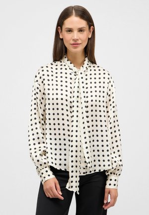 Femme portant un chemisier blanc à pois noirs, manches longues, avec un détail de nouage au col, associé à un pantalon noir.