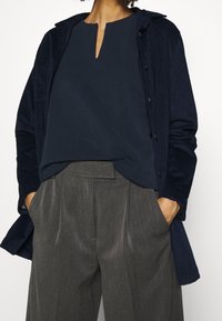 Mörkblå kordjacka med huva över en mörkblå topp med nyckelhålsutskärning, kombinerat med grå höga byxor med veck.