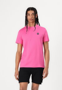 Lyserød bomulds-t-shirt med korte ærmer, prydet med et lille sort logo-patch på brystet. Bæres sammen med sorte shorts for et afslappet look.