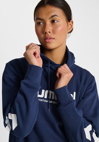 Frau, die die Kapuze eines navyblauen Sportpullovers mit weißem Text und Pfeilgrafiken an den Ärmeln anpasst und zur Seite schaut.