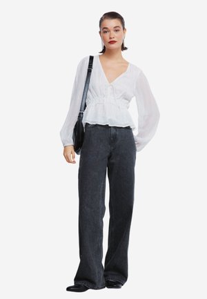 Donna in piedi con una camicia bianca a maniche lunghe, jeans neri a vita alta e gamba larga, scarpe nere e una borsa a spalla nera.