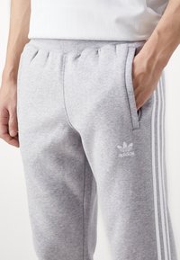 Grå sweatpants med elastisk midja, sidfickor, tre vita ränder längs sidorna och en liten vit Adidas-logotyp på låret.