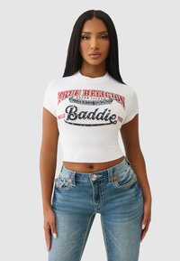 Biały krótki t-shirt z wielokolorowym nadrukiem tekstu "True Religion" oraz "Baddie", zestawiony z jasnoniebieskimi dżinsami z akcentującym przeszyciem.