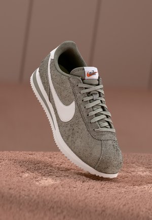 Zapatilla de Nike en ante verde oliva con el logotipo de swoosh blanco, parte superior texturizada y suela de goma de color claro, con cordones y detalles en gris.