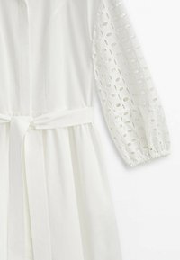 Robe blanche avec des manches longues et un motif découpé circulaire, une taille ceinturée et une silhouette fluide, confectionnée dans un tissu léger.