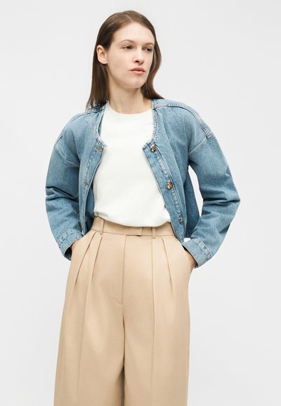 Woman wearing a light blue denim jacket over a white shirt and beige high-waist pleated trousers, standing with hands in pockets.