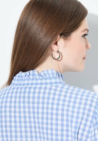 Camicia a quadri blu e bianchi con colletto arricciato, caratterizzata da una texture morbida e una vestibilità rilassata. Sono indossati orecchini a cerchio argentati.