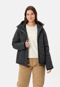 Doudoune noire avec capuche, dotée de fermetures à pression et à glissière, poignets élastiques et texture matelassée. Portée par-dessus un haut de couleur claire.