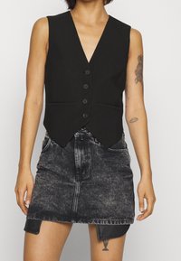 Person med ärmlös svart knäppt väst och blek svart jeanskjol med synliga tatueringar på armar och lår.