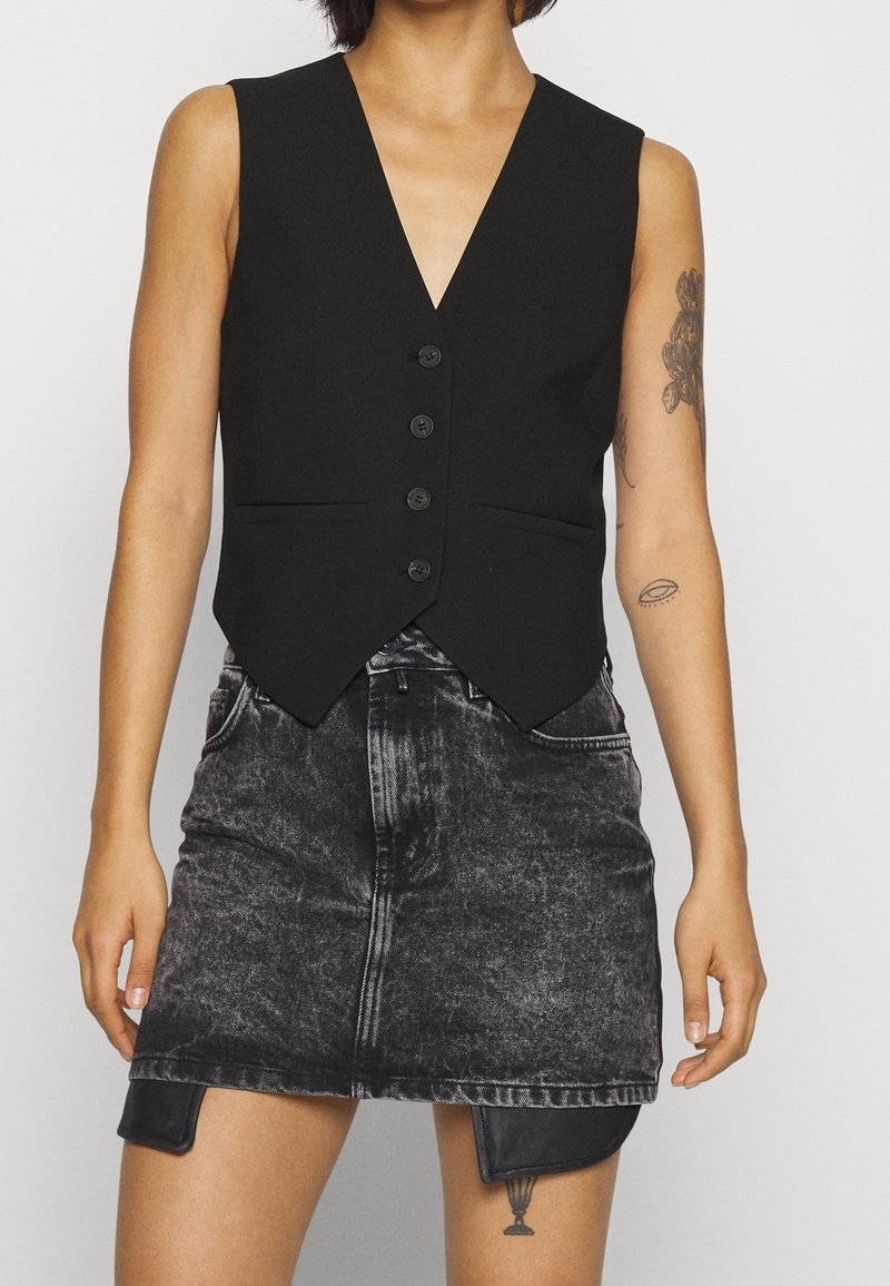 Person med ärmlös svart knäppt väst och blek svart jeanskjol med synliga tatueringar på armar och lår.