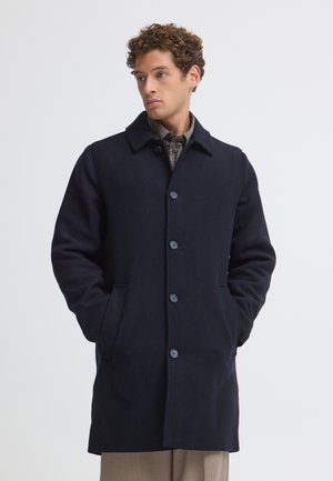 Jeune homme aux cheveux bouclés portant un manteau noir boutonné et un pantalon beige, mains dans les poches, regardant sur le côté sur un fond uni.