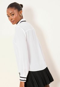 Fehér hosszú ujjú ing fekete gallérral és mandzsettával, fekete redős szoknyával kombinálva. Simább anyag, gombdíszítéssel a mandzsettán.