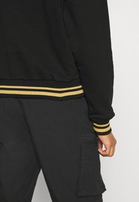 Sudadera negra con puños acanalados amarillos y negros. La tela parece suave y hay un bolsillo en el lado de los pantalones cargo negros.