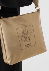 Metalen gouden handtas met een gladde textuur en een ritssluiting. De voorkant toont een minimalistische zwarte lijntekening en logo.