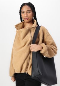 Tan fleece jacket with a high collar and dual zippers, paired with a black leather tote bag, featuring a smooth texture and rectangular shape.