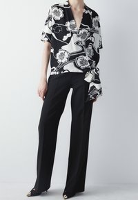 Blusa floreale in bianco e nero con design avvolgente, maniche corte e dettaglio annodato, abbinata a pantaloni neri su misura e scarpe a punta.