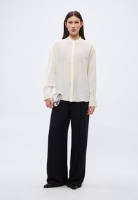 Krémfehér blúz gombos elejjel és hosszú ujjakkal, széles fekete nadrággal és egy kis ezüst színű borítéktáskával, egyszínű háttér előtt.