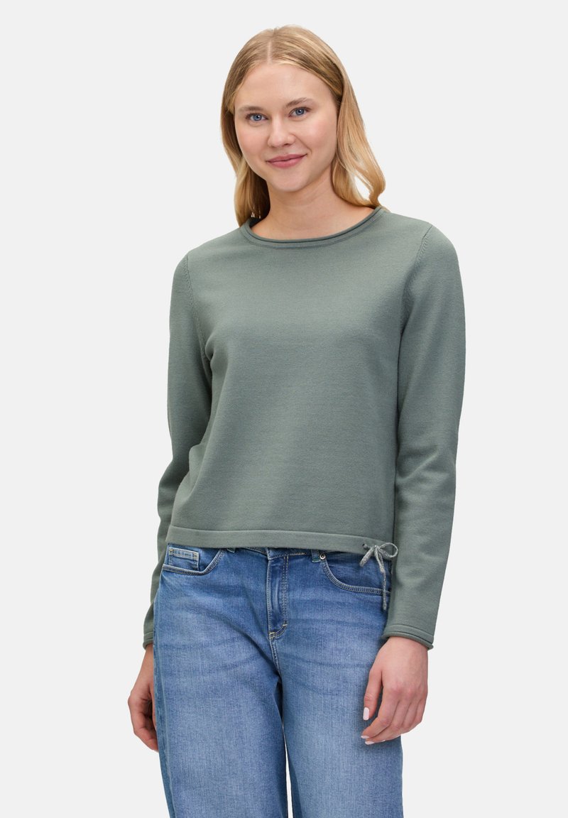 Grüner, langärmeliger Pullover mit Rundhals und einer Kordel am Saum. Strukturierter Stoff, kombiniert mit hellblauen Jeans.