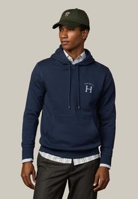 Sudadera azul marino con bolsillo frontal, cordones y logo de Hackett. Lleva puesta sobre una camisa de rayas con cuello y combinada con pantalones grises. Gorra verde.