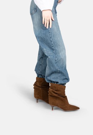 Legs wearing loose blue jeans tucked into brown suede slouchy ankle boots with kitten heels against a white background.